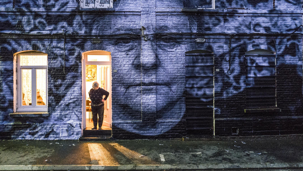 A woman standing in her doorway with a huge print of her own face across the house wall. 