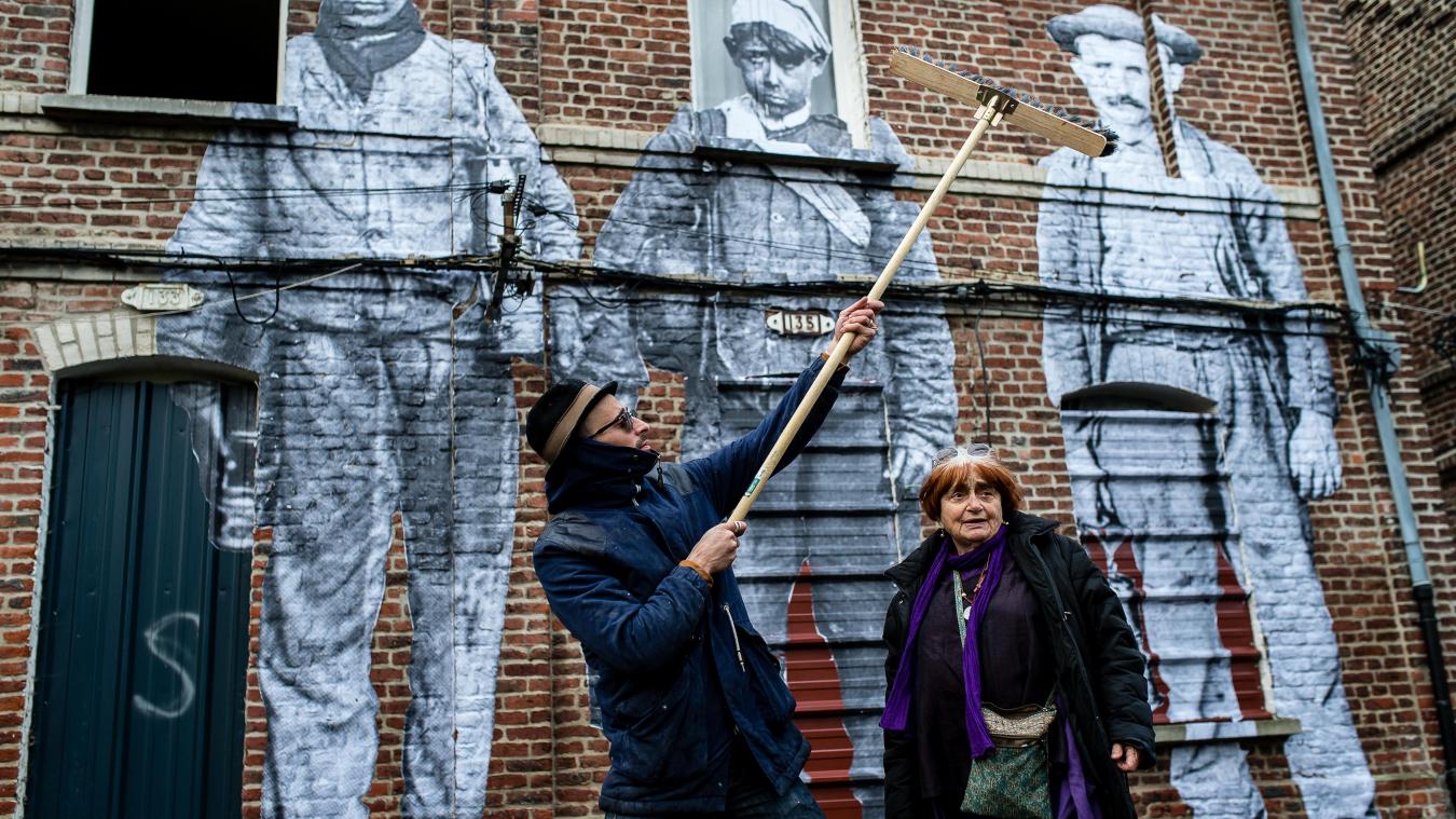 JR and Agnès Varda in front of three prints of workers on a house wall. 