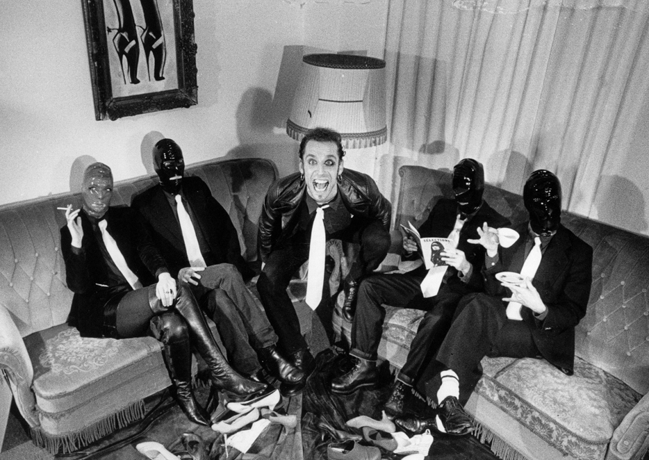Black and white photo of Rosto with four people in BDSM masks grouped around him on two sofas.