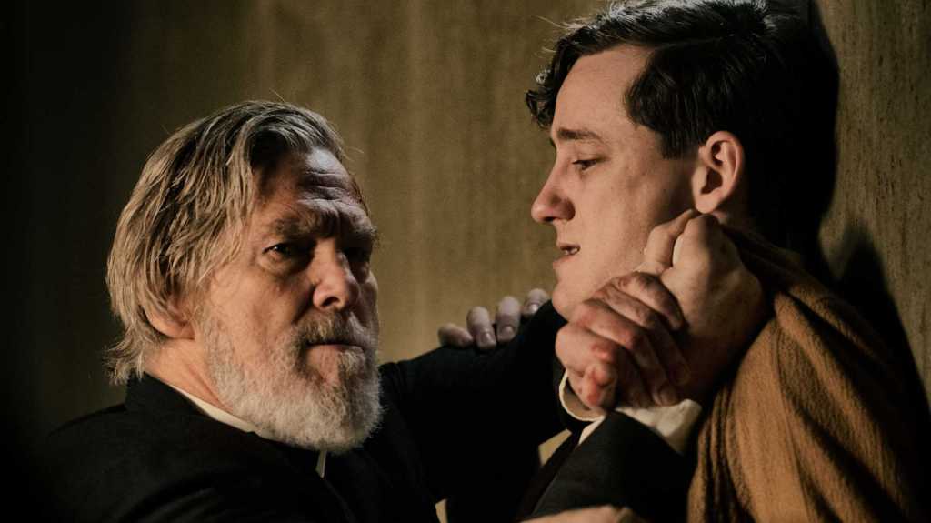 A catholic priest (Jeff Bridges) pushing a young man (Lewis Pullman) against a wall. 