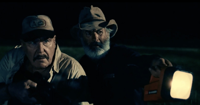 A police man and a farmer standing behind a car, pointing a big flashlight at something.