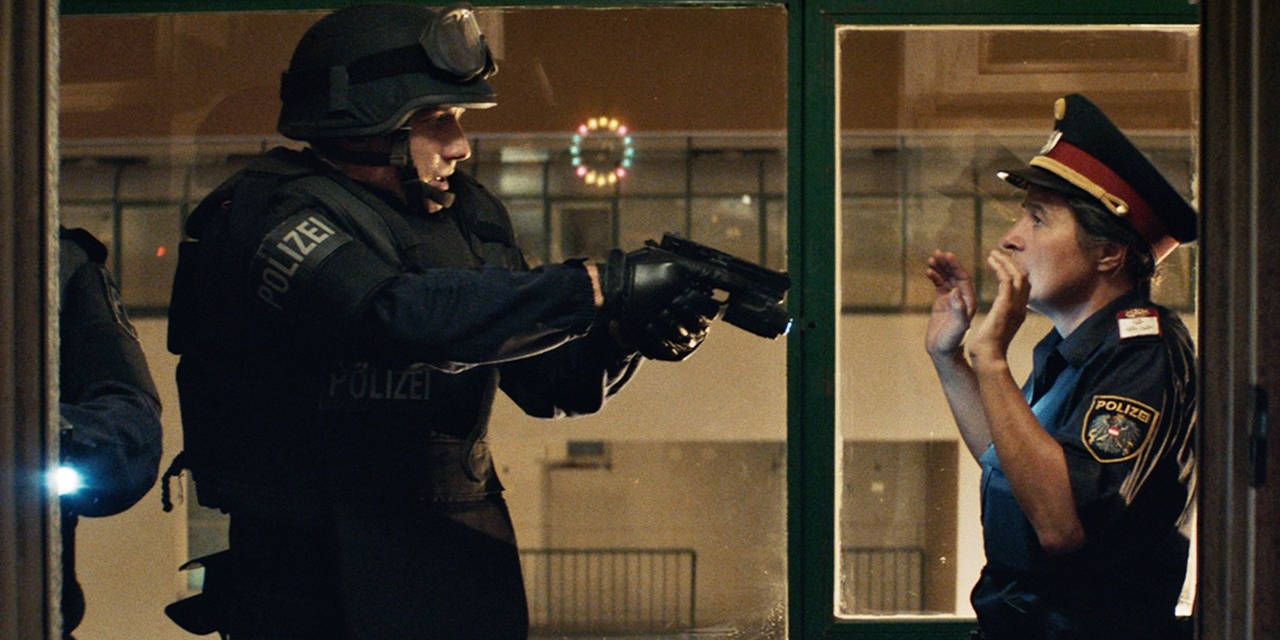 A special forces police officer (Laurence Rupp) in riot gear pointing a gun at a police officer in uniform (Maria Hofstätter). 
