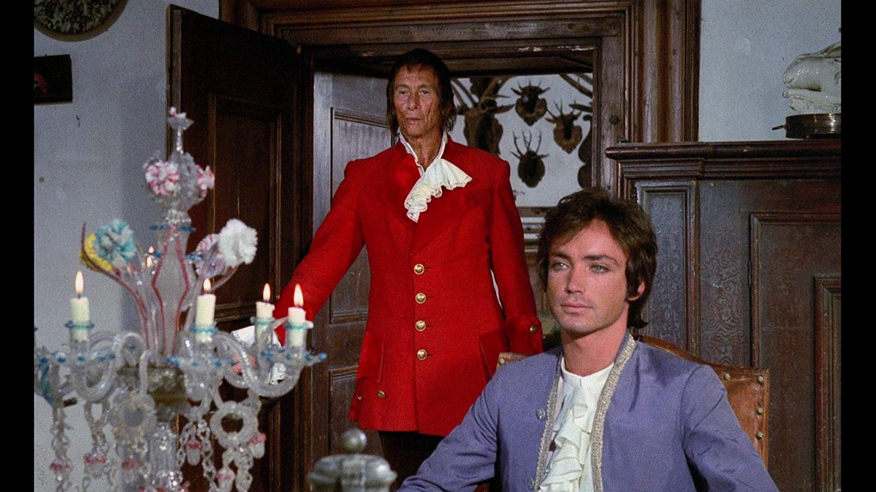 A young man (Udo Kier) sits at the table as an older man (Reggie Nalder) enters the room behind him.