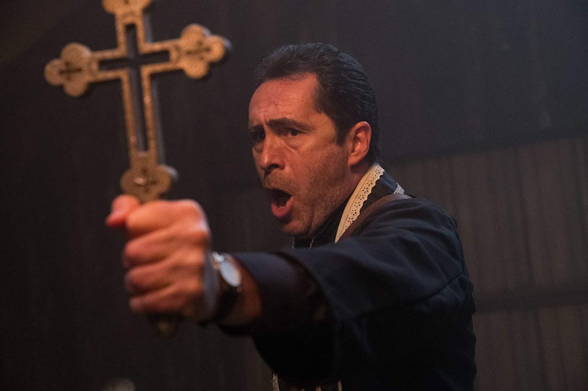 A priest (Demián Bichir) holding out a big cross against something while speaking.