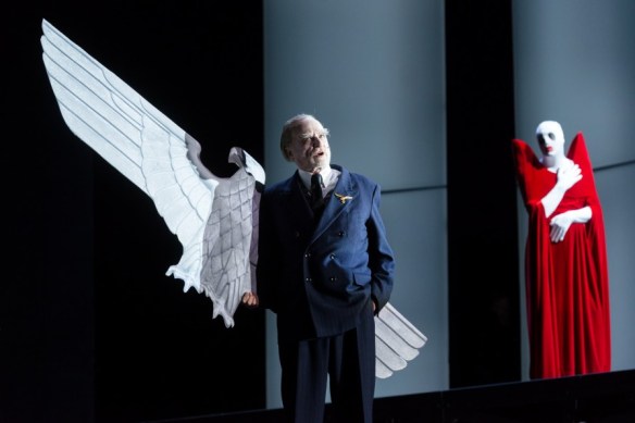 A man in a suit standing in front of a stylized eagle, behind him a figure in a red dress with a white face.