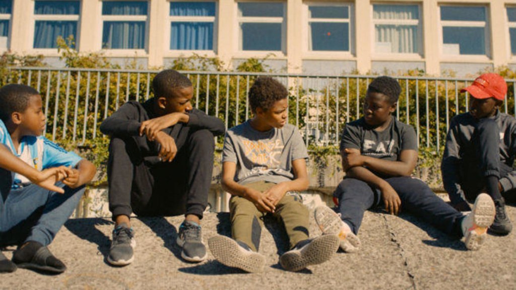 A group of kids, among them Issa (Issa Perica) sitting on the sidewalk. 