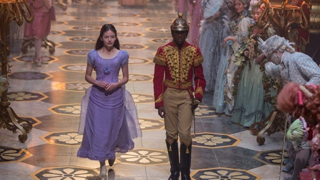 A young girl (Mackenzie Foy) and a soldier (Jayden Fowora-Knight) walk through a crowd of courtiers.