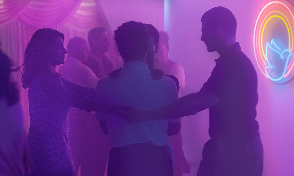 Joana (Dira Paes) and Danilo (Julio Machado) encircling a hugging couple with their arms in dimly lit room.