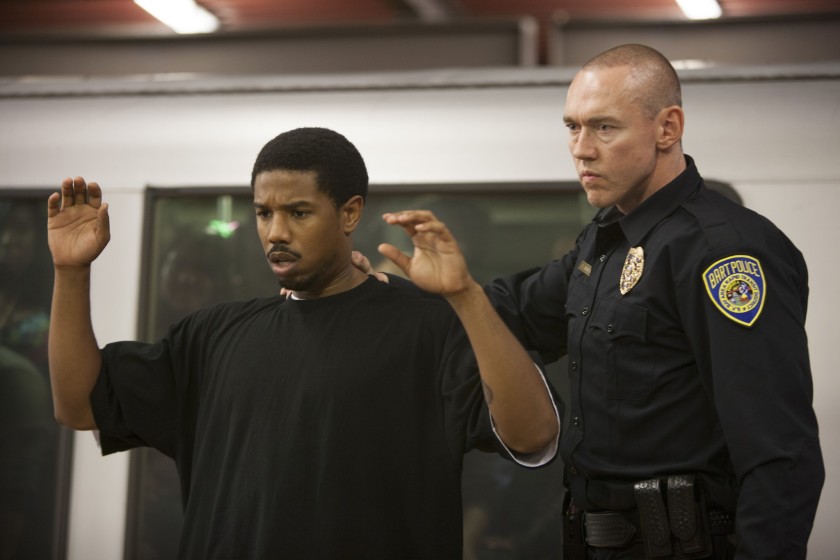 Oscar Grant (Michael B. Jordan) standing with his arms raised and his back turned to a police officer (Kevin Durand). 
