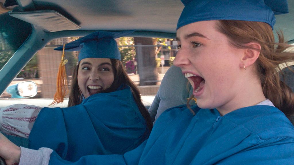 Molly (Beanie Feldstein) and Amy (Kaitlyn Denver) in their graduation caps and gowns, driving while screaming.