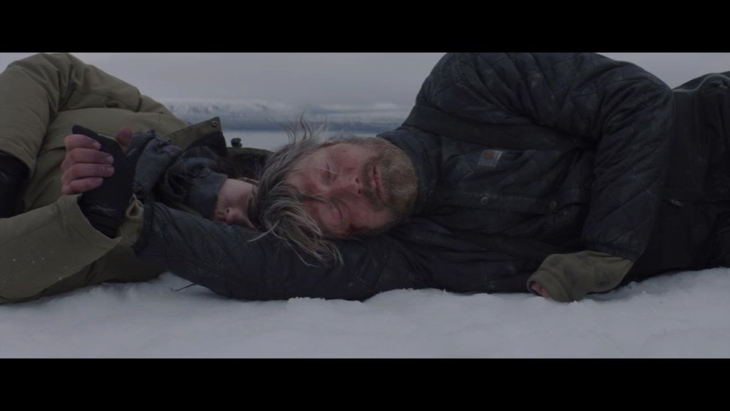 Overgård (Mads Mikkelsen) holding the hand of a young woman (Maria Thelma Smáradóttir) as they both lie in the snow.