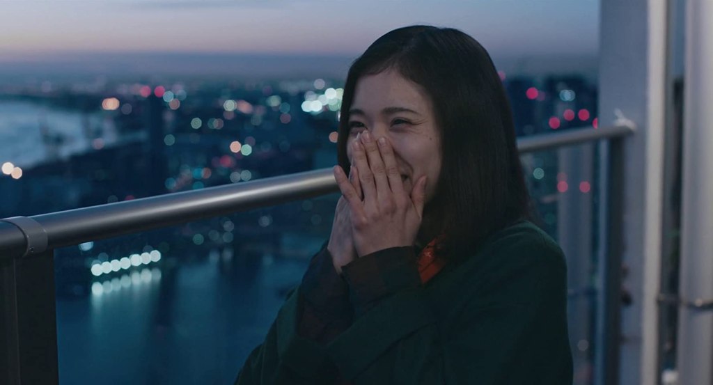 Yoshika (Mayu Matsuoka) smiling on a balcony overlooking the city.
