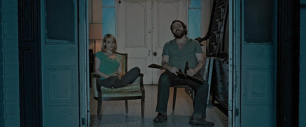 Abby (Brea Grant) and Hank (Jeremy Gardner) sitting in the open door of their house. Hank is cradling a shotgun. 