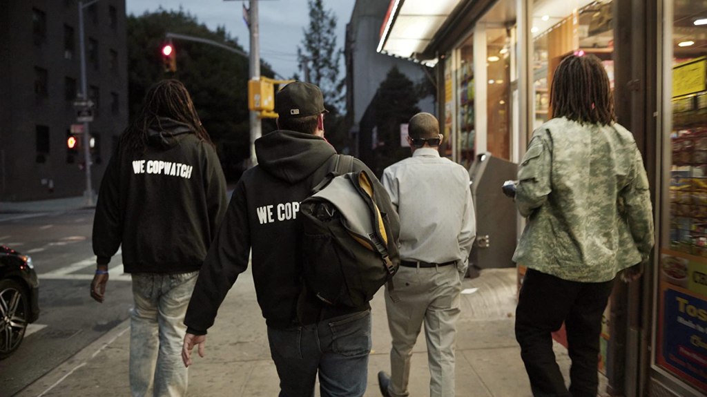 A group of men walking through a street, two are wearing black hoodies with the slogan "We Copwatch".
