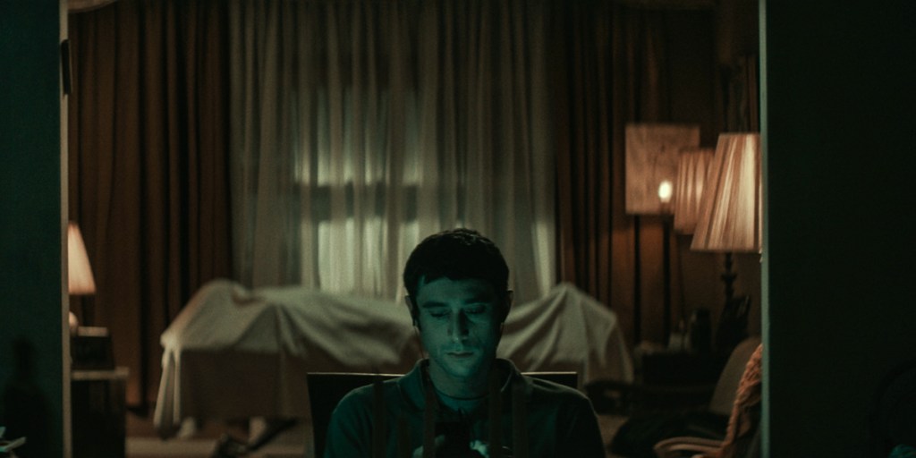 Yakov (Dave Davis) watching something on his cell phone with the corpse in the background. 