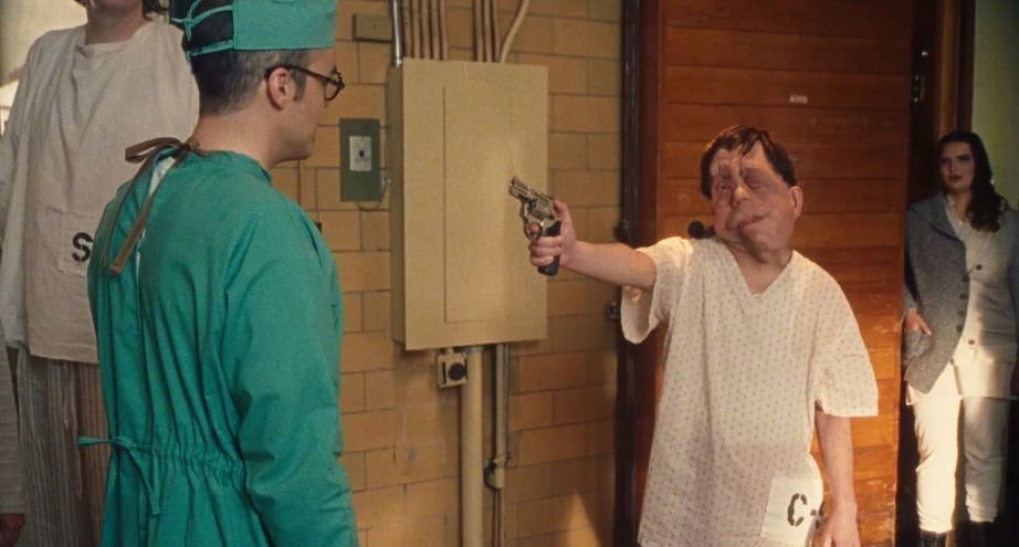 Rosenthal (Adam Pearson) wearing a hospital gown and pointing a gun at a doctor.