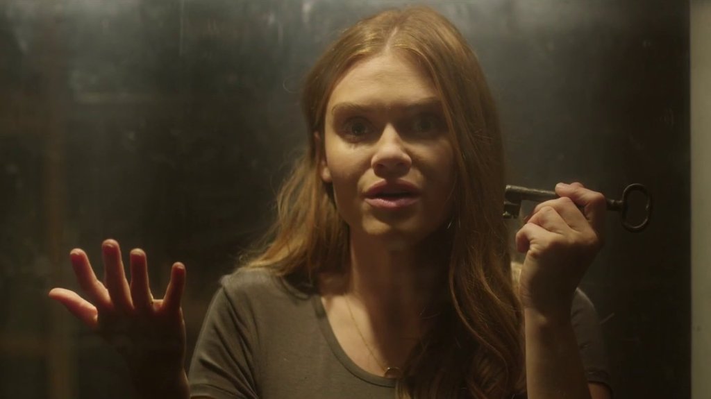 Erin (Holland Roden) inside a glass tank with a puzzled expression and a key in her hand.