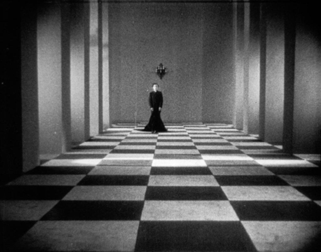 The clergyman (Alex Allin) walking through a hallway with checkered tiles. 