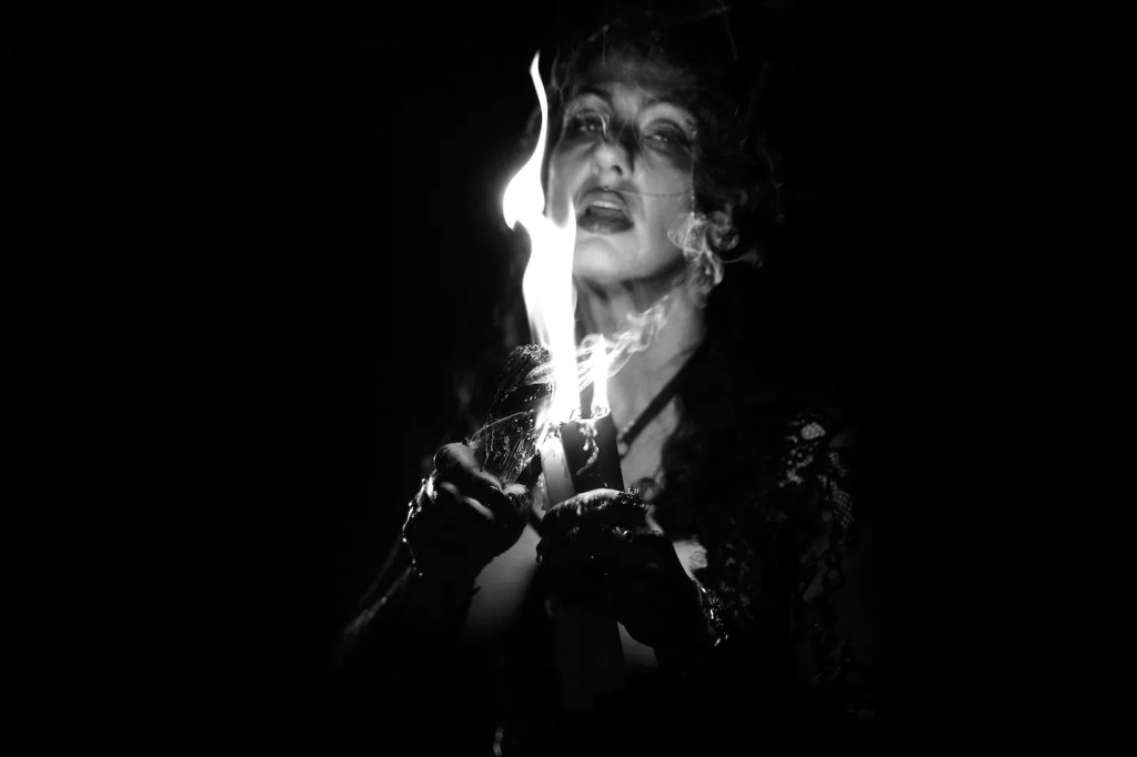 Black and white image: The High Priestess (Sadie Lune) lighting a candle.