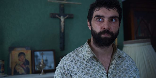Tomas (Alec Secareanu) looking disheveled. A big cross can be seen behind him on the wall.