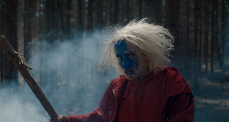 Eve (Lucie Debay) with wild hair, her face covered in blue color, holding up a branch as a weapon.