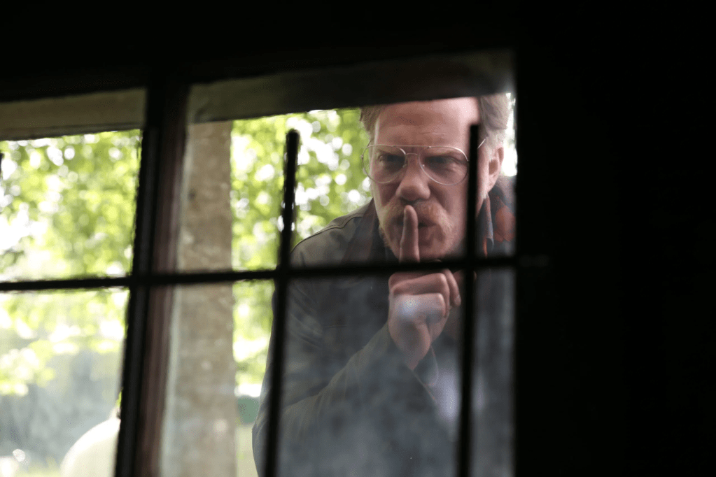 The man (Marc Menchaca) looking in a window, making a shushing gesture.