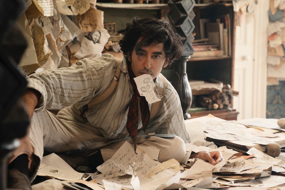 David (Dev Patel) lying in a pile of paper, one of the pieces in his mouth. 