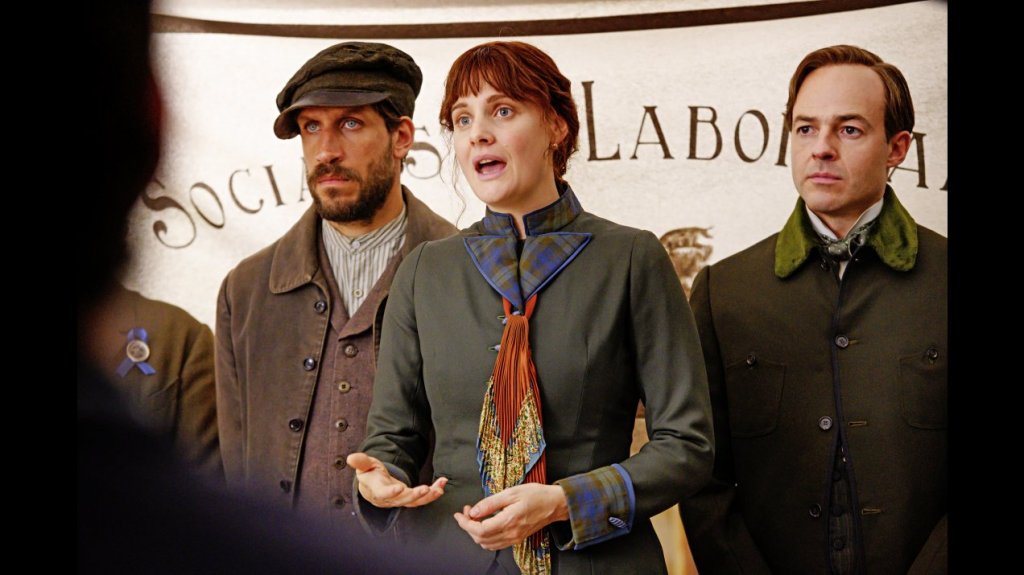 Eleanor Marx (Romola Garai) giving a speech, Edward Aveling (Patrick Kennedy) standing behind her.