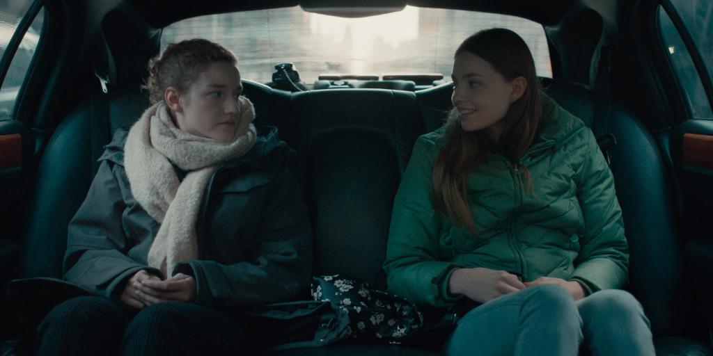 Jane (Julia Garner) driving with Sienna (Kristine Froseth) in the back of a fancy car.