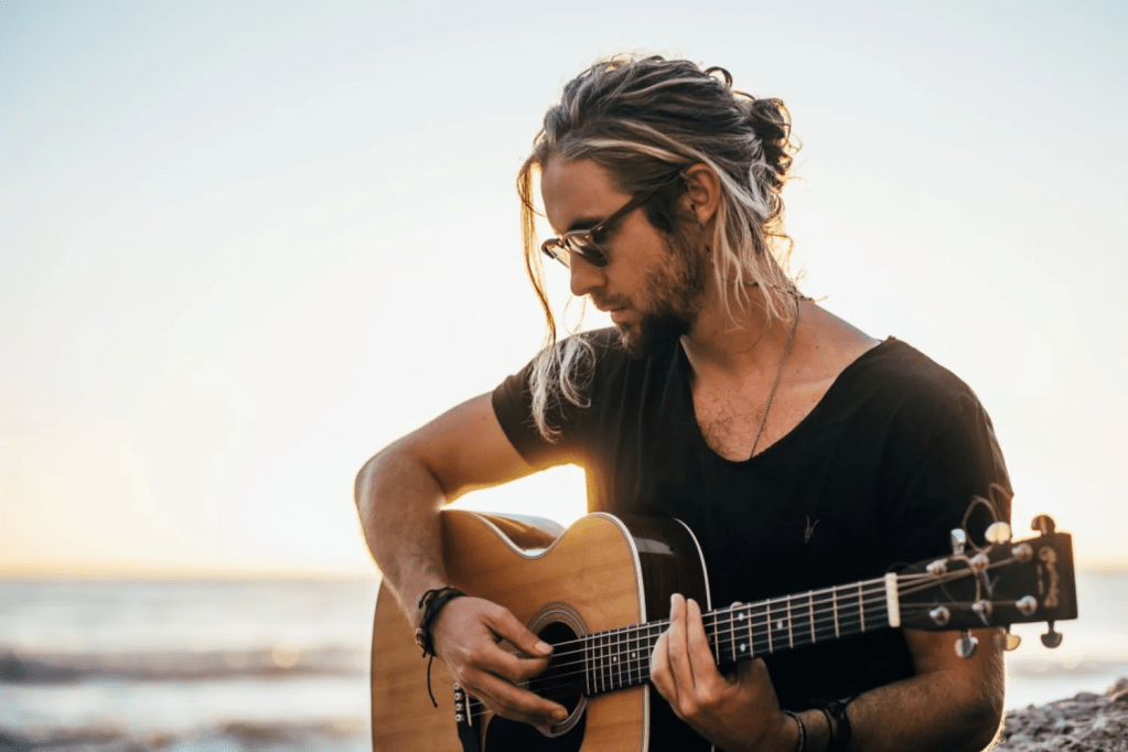 Jeremy Loops playing his guitar on a beach in the sunset.