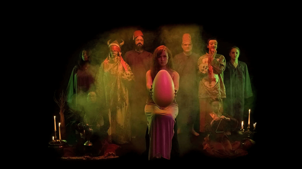 Karen (Holly Bonney) as the center of a surreal tableau, holding out a giant egg.