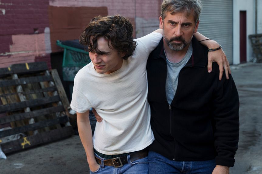 David (Steve Carell) half-carrying Nic (Timothée Chalamet) out of a run-down alley. Nic is crying.