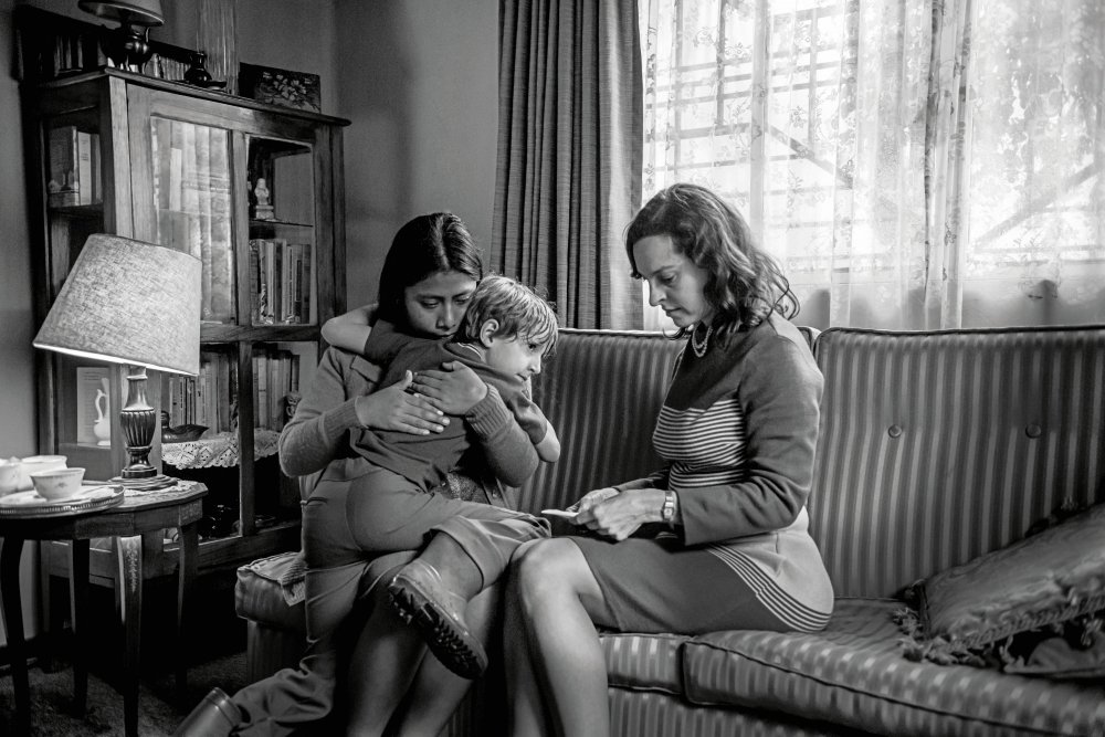 Cleo (Yalitza Aparicio) sitting next to Sra Sofía (Marina de Tavira) on the sofa, hugging Sofía's son Pepe (Marco Graf).