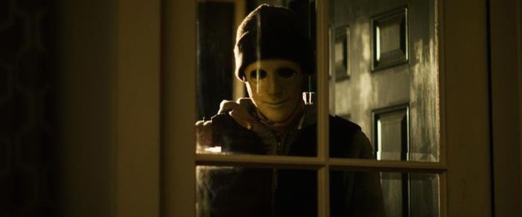 A masked man (John Gallagher Jr.) looking through a glass door into the house.