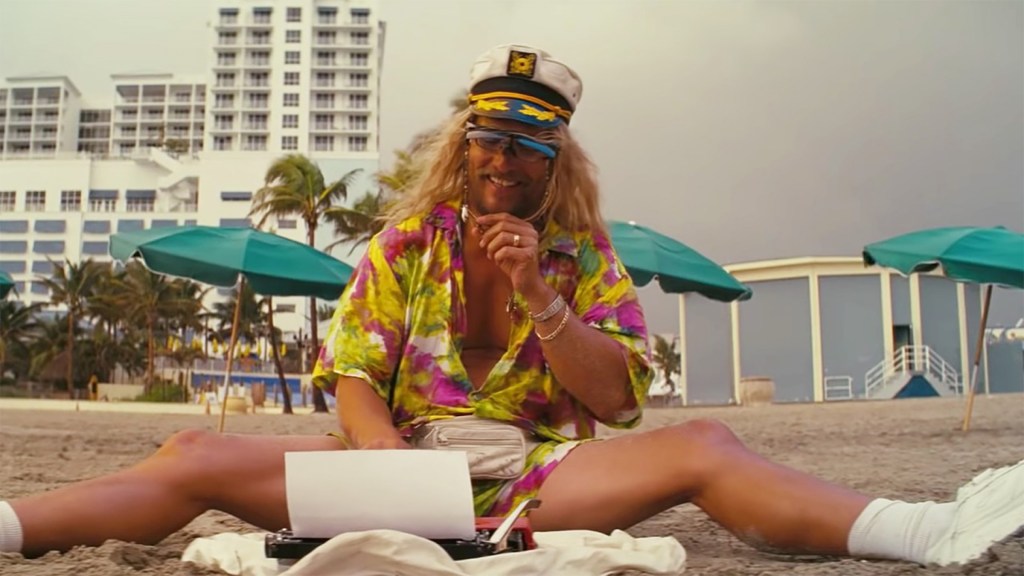 Moondog (Matthew McConaughey) sitting on the beach in a floral dress and a captain's hat with a typewriter in front of him.
