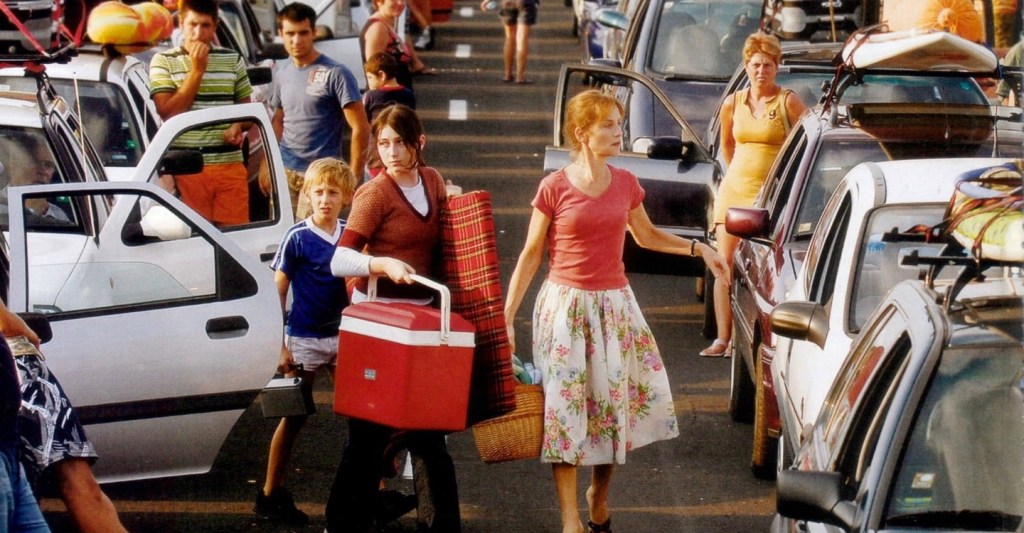 Marthe (Isabelle Huppert) leading her two younger children Marion (Madeleine Budd) and Julien (Kacey Mottet Klein) across the highway during a traffic jam with picknick gear.