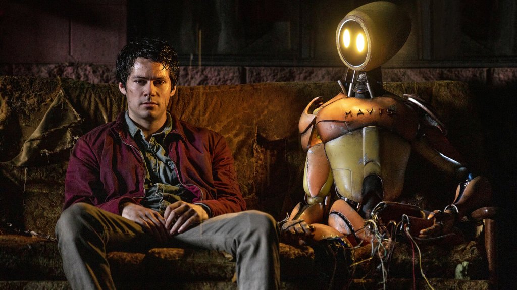 Joel (Dylan O'Brien) sitting on a desolate sofa next to a robot, looking despondent.