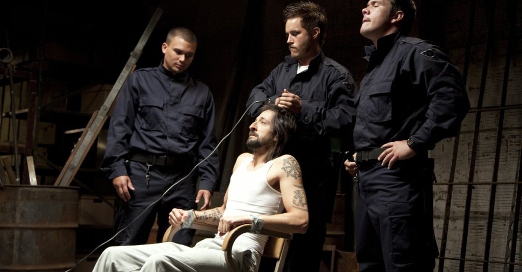 "Guard" Helweg (Travis Fimmel) shaving Travis' (Adrien Brody) head while he is tied to a chair.