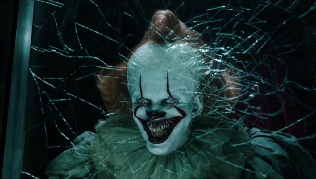 Pennywise (Bill Skarsgård) smiling behind a fractured glass wall, showing a mouth full of sharp teeth.