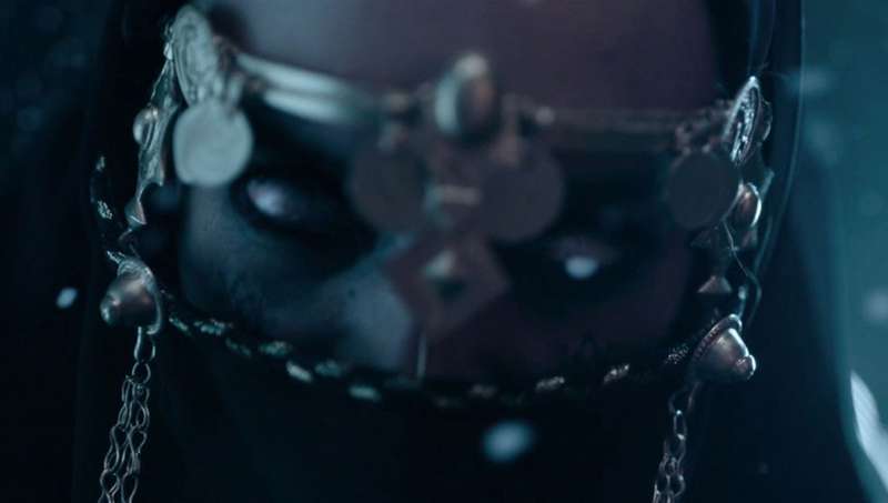 A close-up of a woman - Kandisha (Mériem Sarolie) - wearing a veil in front of her face, her eyes an unnatural shade of silver or white without pupils.