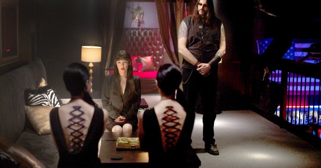 Mary (Katharine Isabelle) meeting with prospective clients. We can only see their backs that have rope threaded directly through the skin as if it was a corset.