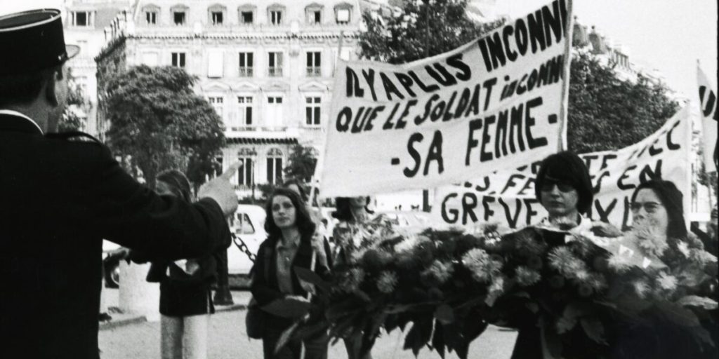 Image of a feminist protest, women are carrying a sign that says "There is someone more unknown than the unknown soldier - his wife" in French. 