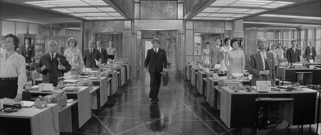 C.R. MacNamara (James Cagney) arriving in his office, with all employees standing at attention while he walks past.