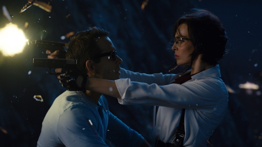 Millie (Jodie Comer) shooting guns with both hands as she sits on Guy's (Ryan Reynolds) lap while he is driving a motorcycle.