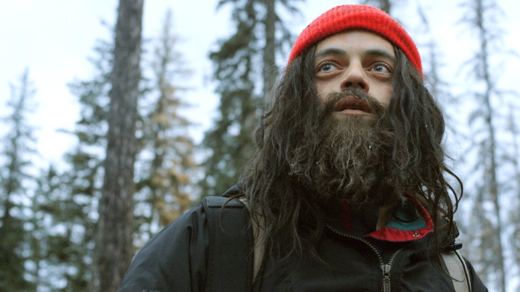 Buster (Rami Malek) with long hair and a long beard, both shaggy, in the forest.
