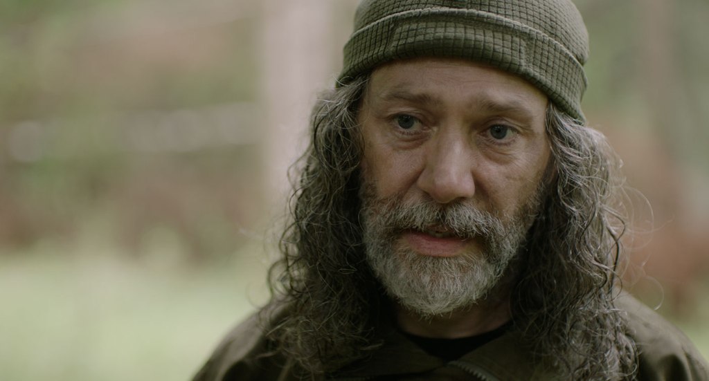 Zach (Reece Shearsmith) with long hair and a beard, wearing a beanie.