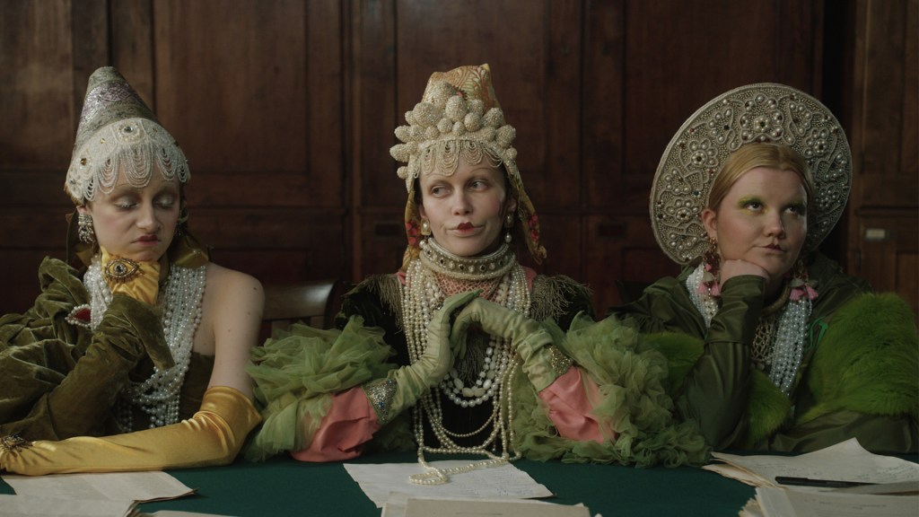 Three women in fancy headdresses and ballgowns sitting at a table looking very bored.