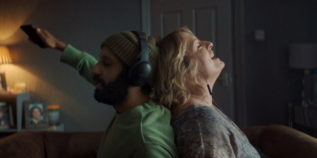 Ali (Adeel Akhtar) and Ava (Claire Rushbrook) sitting back to back as they both listen to different music on their headphones.