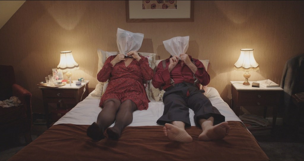 A couple lying on a bed together with plastic bags over their heads.