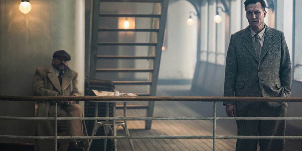 Josef Bartok (Oliver Masucci) standing on the deck of the cruise ship, talking to another man (Samuel Finzi). 